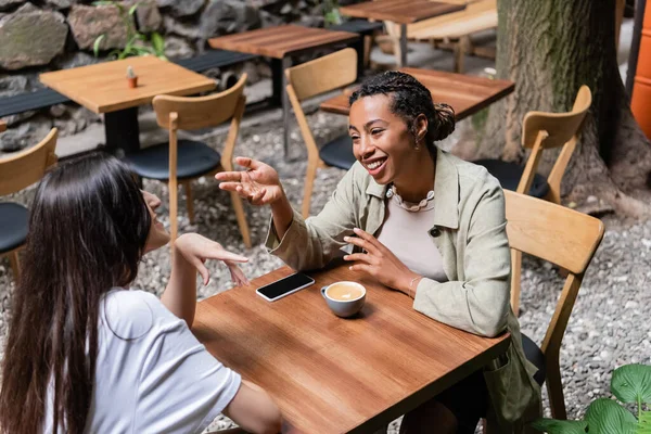 African american woman talking to friend near coffee and smartphone on terrace of cafe — Stockfoto