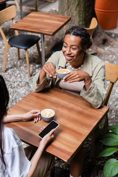 High angle view of cheerful african american woman holding cup of coffee near friend with cellphone on terrace of cafe — Stockfoto