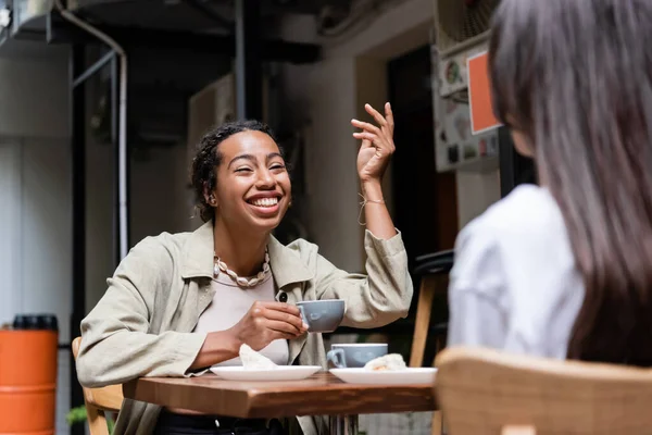 African american woman holding coffee cup and talking to blurred girlfriend on terrace of cafe — Stock Photo