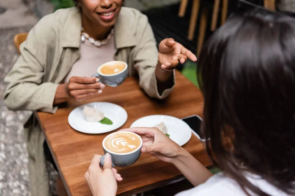Розмита жінка тримає чашку капучино біля афроамериканського друга у відкритому кафе — стокове фото
