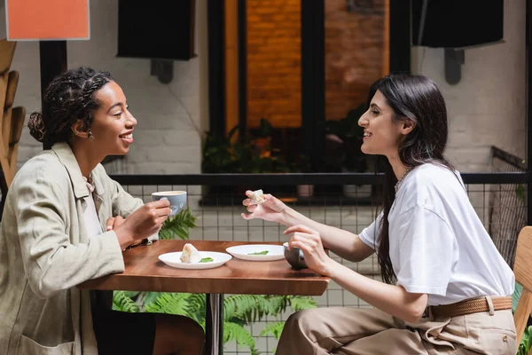 Side view of multiethnic friends talking near coffee and desserts in outdoor cafe — Stock Photo