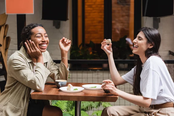 Laughing multiethnic girlfriends talking near coffee and desserts on cafe terrace — Stock Photo