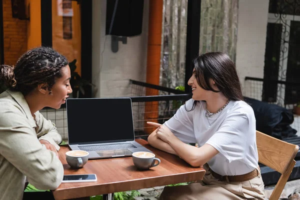 Side view of smiling woman pointing at laptop near coffee and african american friend in cafe — Foto stock