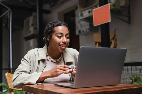 African american woman holding cup and looking at laptop on terrace of cafe — Stockfoto