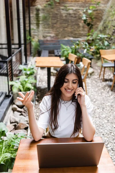 Smiling freelancer talking on smartphone near laptop on terrace of cafe — Stock Photo