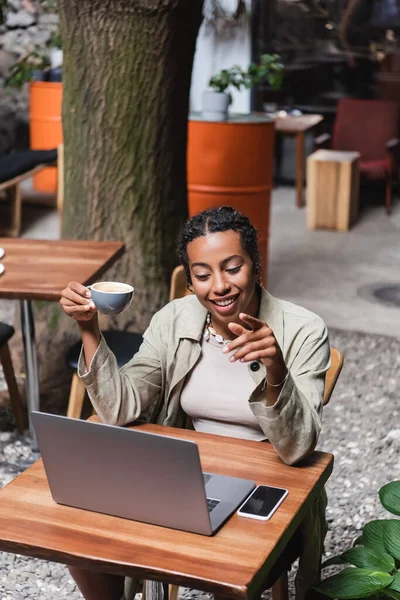 Positive african american woman holding cup and pointing with finger during video call on laptop on terrace — Foto stock