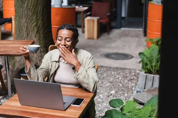 Laughing african american woman holding cup of coffee near devices in outdoor cafe — Photo de stock