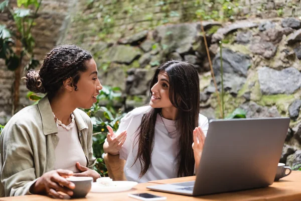 Interracial girlfriends talking near gadgets and coffee in outdoor cafe - foto de stock