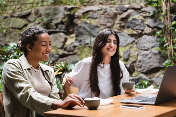 Smiling interracial girlfriends with cups of coffee looking at laptop in outdoor cafe — Photo de stock