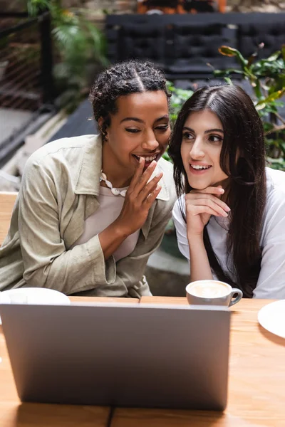 African american woman telling secret to friend near coffee and laptop in outdoor cafe — Fotografia de Stock