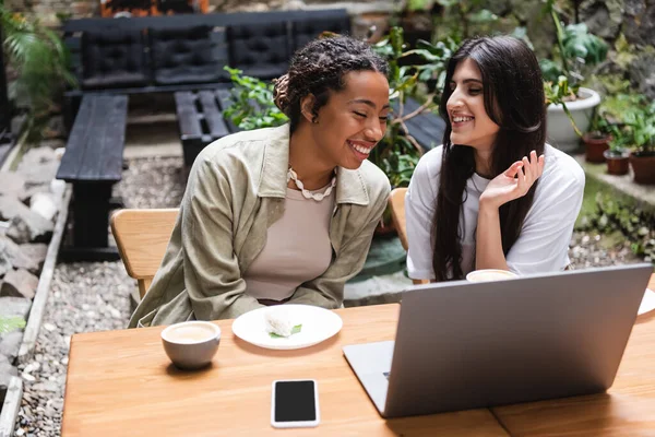 Cheerful multiethnic friends spending time near gadgets and coffee in outdoor cafe — Stock Photo