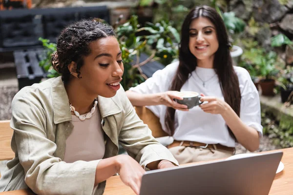 Smiling african american woman using laptop near blurred friend with coffee in outdoor cafe - foto de stock