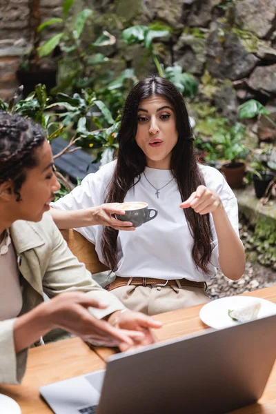 Shocked woman holding coffee and pointing at laptop near blurred african american friend in outdoor cafe — Stock Photo