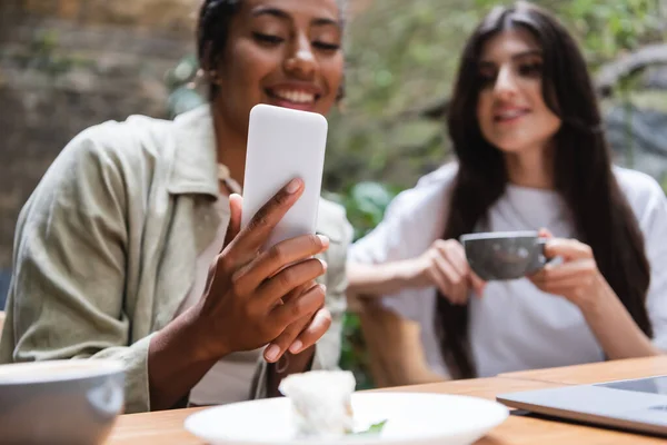 Смартфон в руке размытой африканской женщины, сидящей рядом с другом с чашкой в кафе на открытом воздухе — стоковое фото