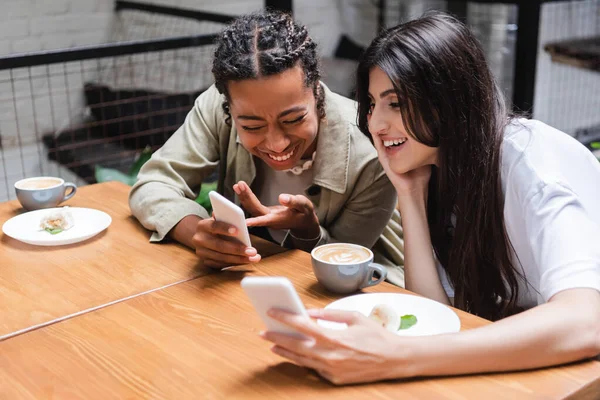 Positive multiethnic girlfriends using smartphones near coffee and desserts in outdoor cafe — Stockfoto