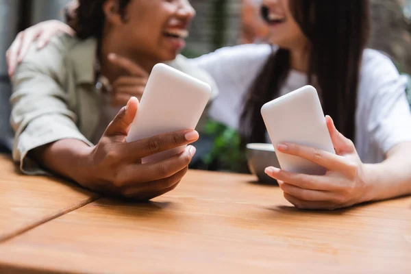 Cropped view of blurred interracial girlfriends holding smartphones in outdoor cafe - foto de stock