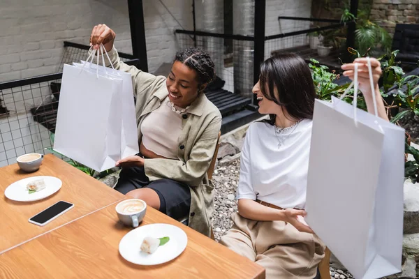 Multiethnic girlfriends holding shopping bags near desserts and coffee on terrace of cafe — Stock Photo