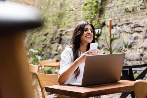 Smiling woman holding smartphone near laptop and looking away in outdoor cafe - foto de stock
