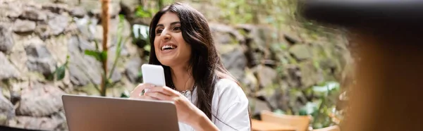 Happy woman holding smartphone near laptop in outdoor cafe, banner - foto de stock