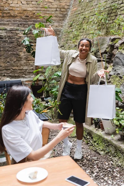 Cheerful african american woman holding shopping bags near friends with cup of coffee in outdoor cafe — Foto stock