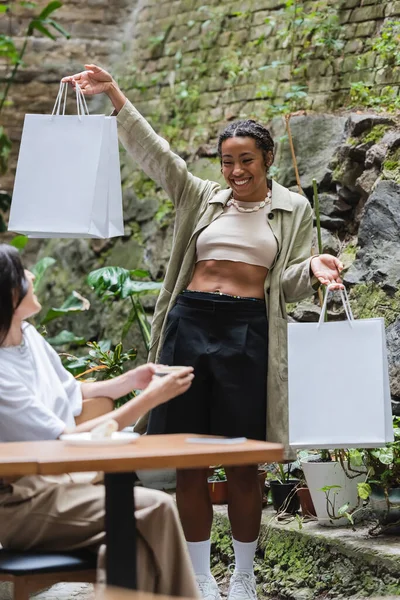 Позитивна афро-американська жінка, яка тримає сумки біля розмитого друга з кавою на терасі. — стокове фото