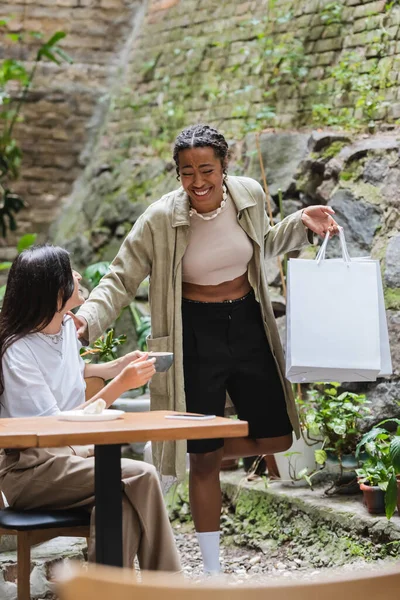 Positive african american woman holding shopping bags near friend with coffee in outdoor cafe — Photo de stock