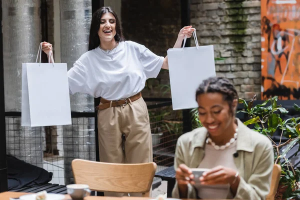 Cheerful woman holding shopping bags near blurred african american friend with coffee in outdoor cafe — Stock Photo