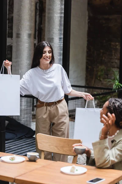 Positive woman holding purchases near african american friend with coffee in outdoor cafe — Fotografia de Stock