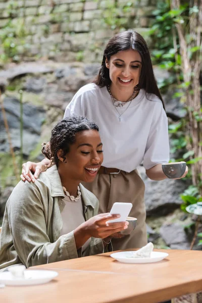 Smiling woman holding cup near african american friend with smartphone in outdoor cafe — Stockfoto