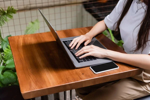 Обрезанный вид женщины с помощью ноутбука рядом со смартфоном в открытом кафе — стоковое фото