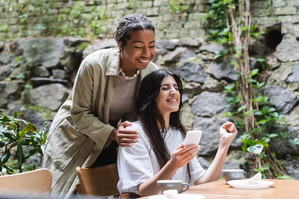Cheerful african american woman standing near friend holding smartphone and showing yes gesture in outdoor cafe — Stock Photo