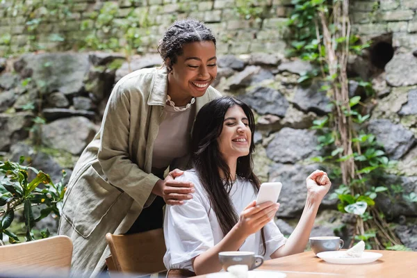 Cheerful african american woman standing near friend holding smartphone and showing yes gesture in outdoor cafe - foto de stock