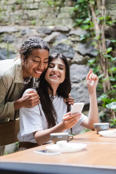 Positive african american woman hugging friend with smartphone near cups of coffee in outdoor cafe — Stockfoto