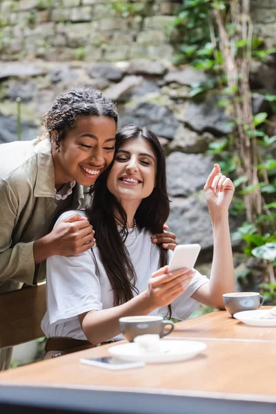 Positive african american woman hugging friend with smartphone near cups of coffee in outdoor cafe — Stock Photo