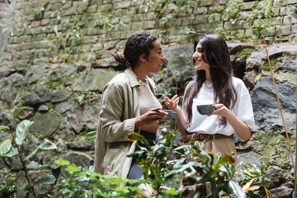 Side view of smiling interracial friends with coffee talking near plants in outdoor cafe — Foto stock