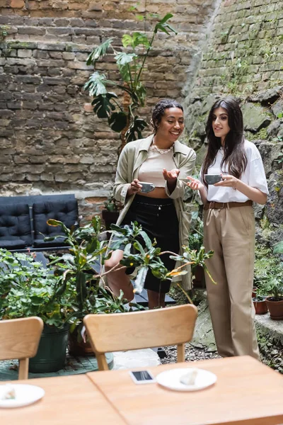 Smiling african american woman holding cup of coffee and talking to friend on terrace of cafe — Stock Photo