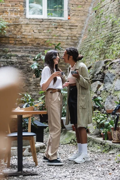 Laughing interracial girlfriends holding cups of coffee in outdoor cafe — Fotografia de Stock