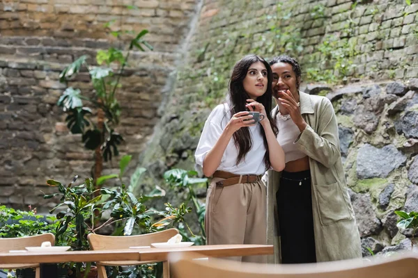 African american woman telling secret to amazed friend with coffee on terrace of cafe - foto de stock