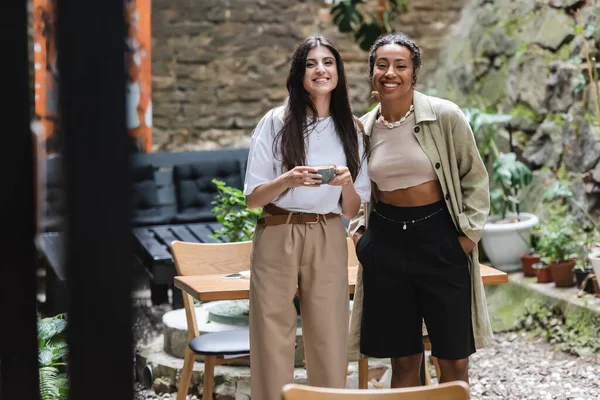 Positive interracial friends with coffee looking at camera on terrace of cafe — Foto stock