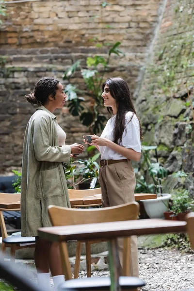 Side view of multiethnic girlfriends holding coffee cups while talking on terrace of cafe — Stock Photo