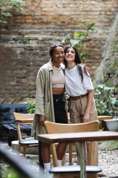 Smiling interracial girlfriends hugging and looking at camera on terrace of cafe — Stock Photo