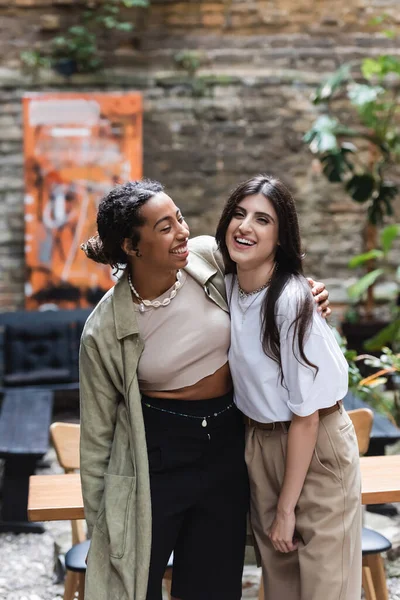 Cheerful african american woman hugging friend on terrace of cafe — Stock Photo