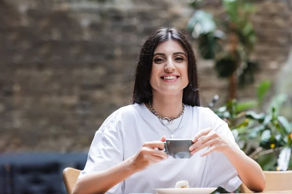Smiling woman holding cup of coffee near dessert on terrace of cafe — Photo de stock