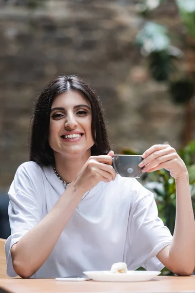 Cheerful brunette woman holding cup of coffee near dessert and smartphone in cafe — Stock Photo