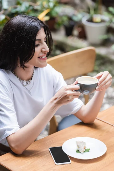 Brunette woman holding cup of cappuccino near dessert and smartphone on terrace of cafe — Stockfoto