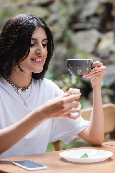 Positive brunette woman holding dessert and coffee near cellphone on terrace of cafe — Foto stock