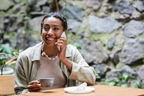 Улыбающаяся африканская американка разговаривает на смартфоне рядом с кофе и десертом на террасе кафе — стоковое фото