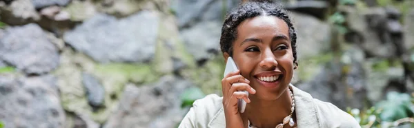 Cheerful african american woman talking on smartphone on terrace of cafe, banner - foto de stock