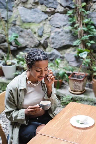 African american woman talking on smartphone and holding cup near dessert in outdoor cafe — Photo de stock