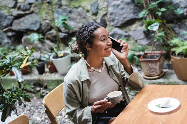 Веселая африканская американка разговаривает по смартфону и держит чашку капучино в открытом кафе — стоковое фото